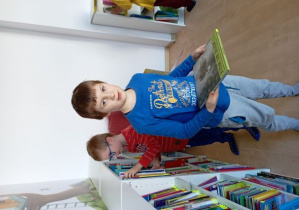 Chłopcy przy regale z książkami. Jeden trzyma w rękach wybraną pozycję, drugi wybiera książkę spośród tych ustawionych na półkach